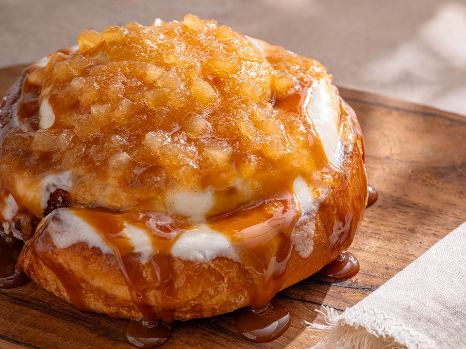 A freshly baked cinnamon roll glazed with cream cheese frosting, topped with spiced apple compote and drizzled with salted caramel.