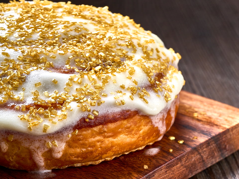 A freshly baked cinnamon roll glazed with lemon cream cheese icing and gilded with luxurious gold sprinkles.