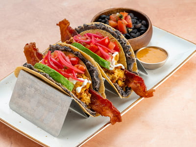 Two wheat-corn tortillas with refried black beans wrapped around crispy corntortilla shells filled with cheesy scrambled cage-free eggs, chorizo and a slice ofcrispy hardwood smoked bacon. Topped with lime crema, housemade pico de gallo,fresh avocado and house-pickled red onions. Served with chipotle queso and a sideof seasoned black beans.