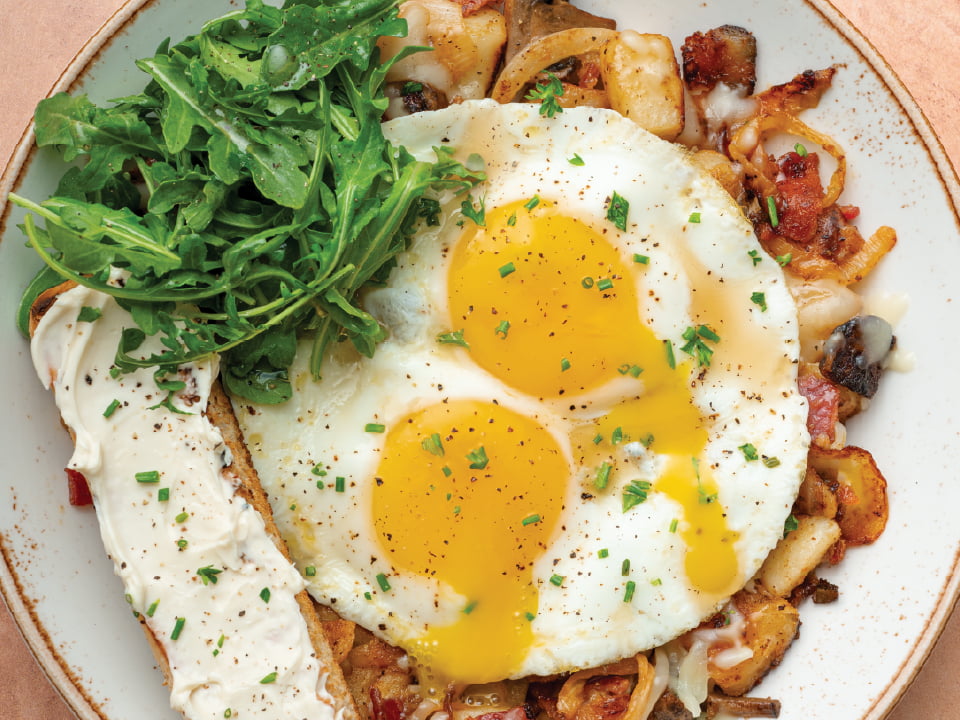 A skillet of crispy breakfast hash topped with melted brie cheese, thick-cut bacon, and a drizzle of truffle oil.